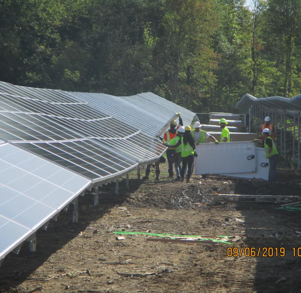 A group of workers in safety gear install solar panels in a field surrounded by trees. The panels are arranged in rows, and the workers are wearing helmets and high-visibility vests. The scene is lit by sunlight, and the date on the image is September 6, 2019.