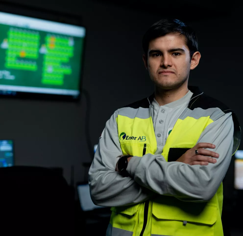 A person wearing a high-visibility vest stands with arms crossed in a control room. Multiple computer screens displaying data and graphics are visible in the background.