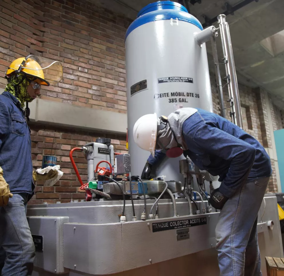 Two workers in protective gear are inspecting equipment in an industrial setting. One worker is standing, holding a can and cloth, while the other is bent over, examining a large metal tank labeled 'ACEITE MOBIL DTE 26 385 GAL.' The setting includes brick walls and control panels.