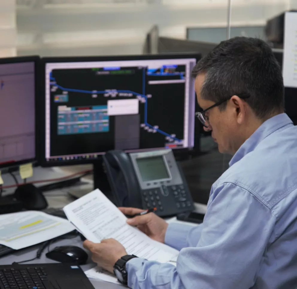A man in a blue shirt sits at a desk with multiple computer monitors displaying data and graphs. He is holding and reading a stack of documents. A telephone is placed on the desk next to the monitors.