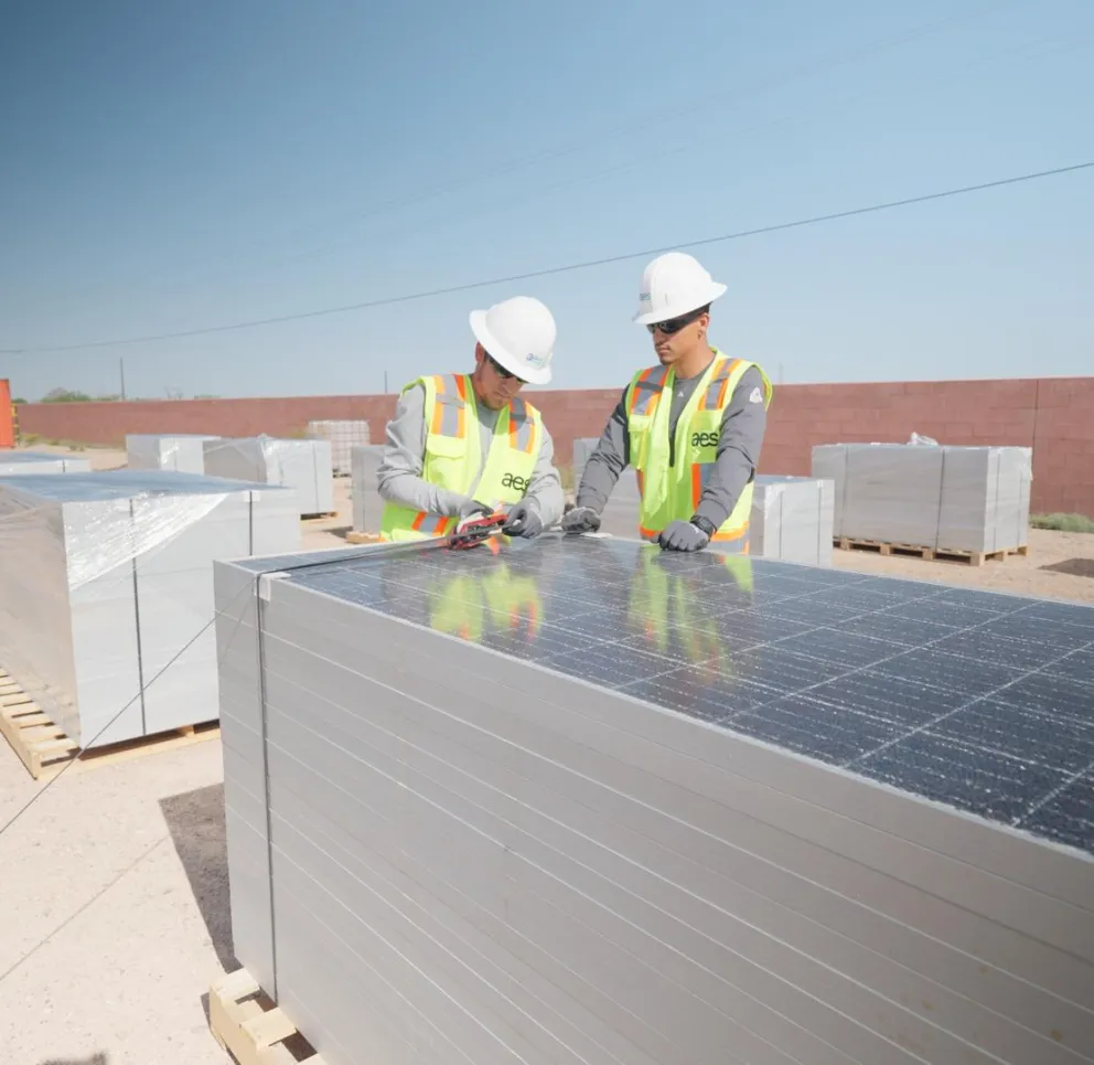 Two workers in safety gear and helmets inspect a stack of solar panels on a pallet in a sunny outdoor area. Other pallets with wrapped panels are visible in the background, along with a shipping container and a brick wall.
