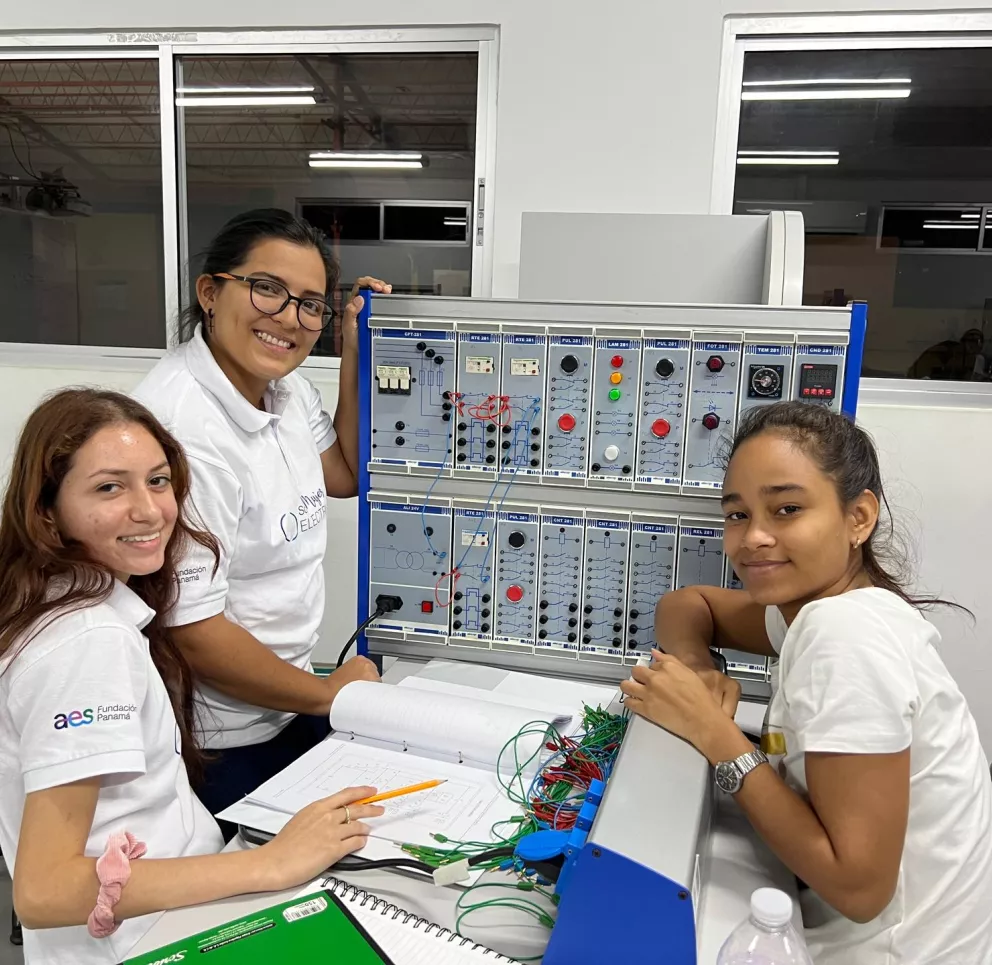 Three young women in white shirts work together on an electrical engineering project in a classroom. They are using a panel with various controls and wires. One woman holds a pencil and open notebook, while the others look at the equipment. They are smiling and appear engaged in their work.