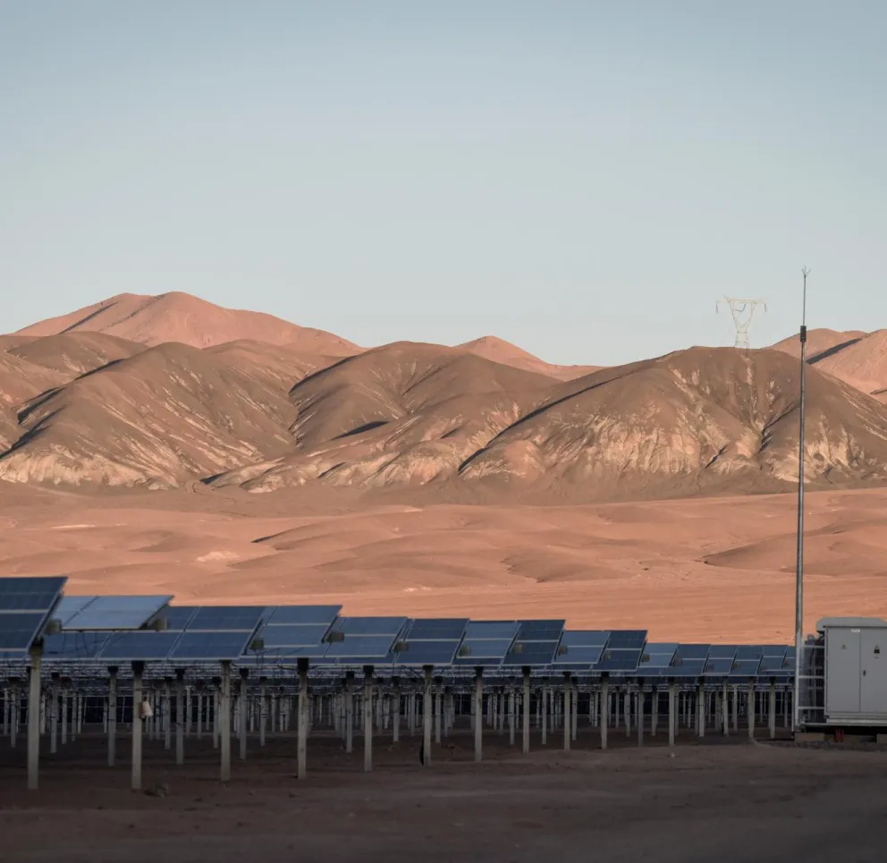 A solar farm with rows of solar panels is set against a backdrop of barren, brown hills under a clear blue sky.