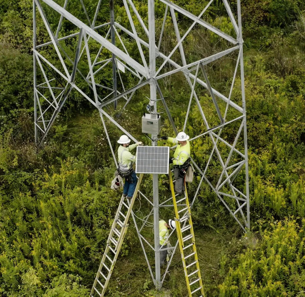 Three workers in safety gear are on ladders installing a solar panel on a metal tower in a lush, green field. One worker is on the ground nearby.