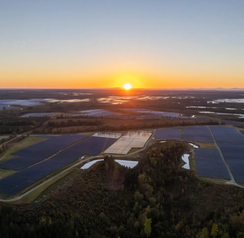 Aerial view of a solar farm at sunset. Rows of solar panels cover large fields, reflecting the orange and yellow hues of the setting sun. The horizon shows a line of distant hills under a clear sky.