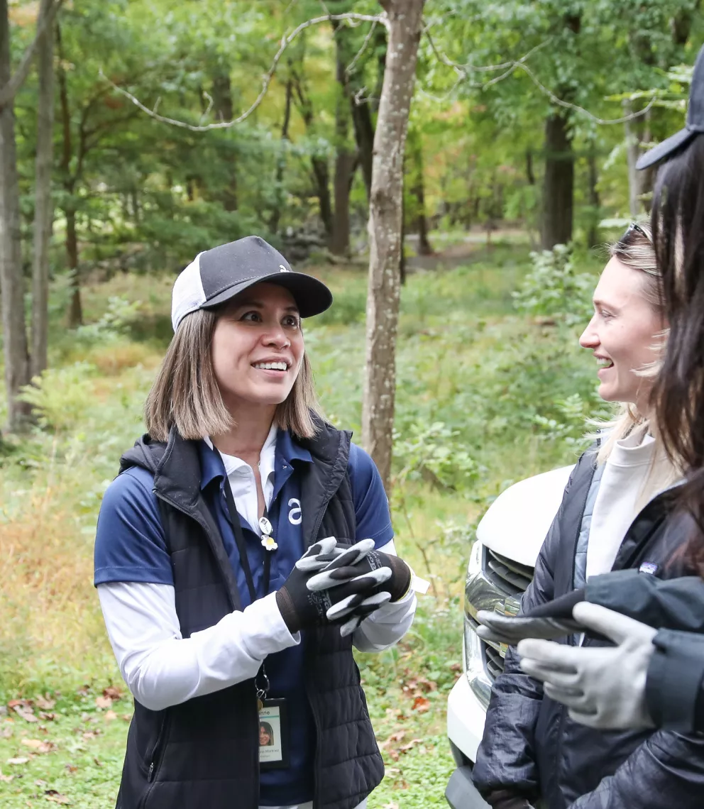 Three people wearing casual outdoor clothing and gloves are smiling and chatting in a forested area. One woman is wearing a baseball cap and holding her hands together.