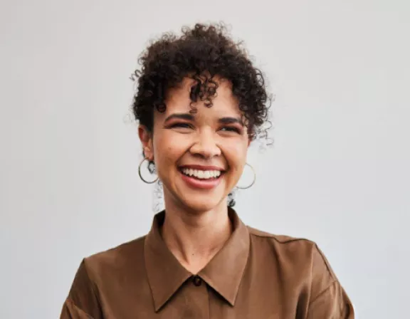 A person with curly hair smiles while wearing a brown shirt and hoop earrings, standing against a plain background.