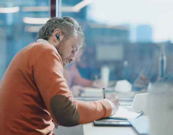 A person with earbuds is sitting at a desk, writing in a notebook. They are wearing an orange sweater with elbow patches. The setting appears to be an office with a blurred background and a laptop on the desk.