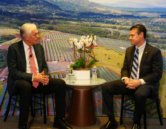 Two men in suits sit facing each other in front of a backdrop depicting a solar farm. A small table with a potted orchid and bottled water is between them.