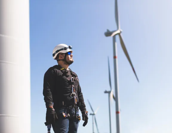 A person wearing a safety harness, gloves, and a white helmet stands outdoors, looking up. They are in front of wind turbines under a clear blue sky.