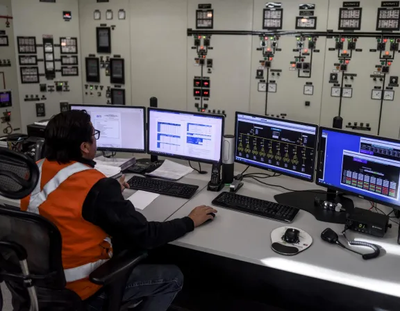 A person wearing an orange safety vest sits at a control room desk with multiple computer monitors displaying data and graphs. The background shows a wall filled with control panels and gauges.
