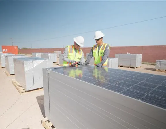 Two workers wearing safety vests and helmets inspect a stack of solar panels on a construction site. The panels are placed on wooden pallets, and more stacks are visible in the background. The site is outdoors with a clear sky.