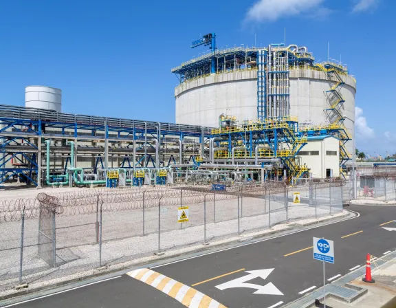 Industrial facility with a large cylindrical storage tank, surrounded by blue and yellow metal structures and pipelines. The area is enclosed by a chain-link fence topped with barbed wire. A road with directional arrows and traffic cones is in the foreground under a clear blue sky.