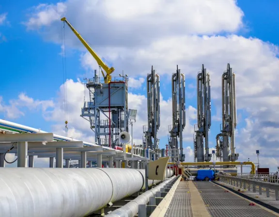 Industrial scene with large pipelines and machinery under a blue sky with clouds. A yellow crane is visible on the left, and several tall, vertical structures are on the right.