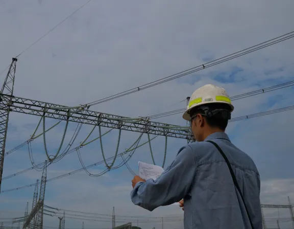A person wearing a hard hat and blue shirt stands outdoors, holding papers and looking at electrical transmission towers and power lines against a cloudy sky.