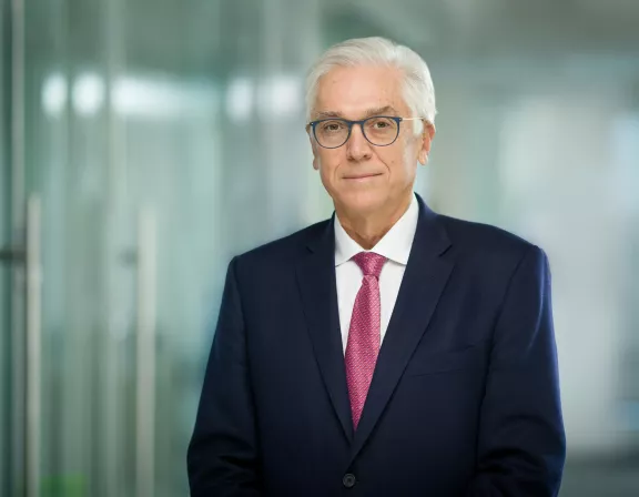 A man with white hair and glasses wearing a navy suit and pink tie stands in a modern office setting with glass walls in the background.