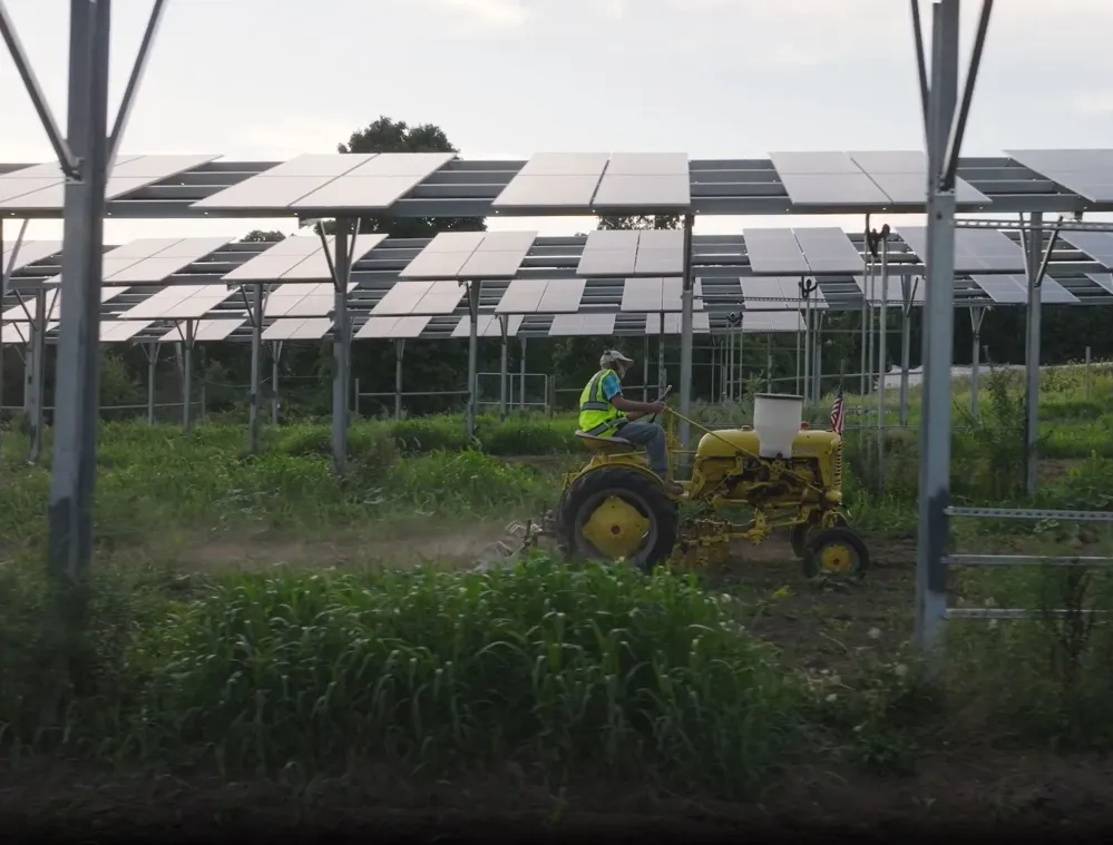 A person wearing a safety vest operates a yellow tractor under a large array of solar panels. The area is surrounded by green vegetation, and the sky is overcast.