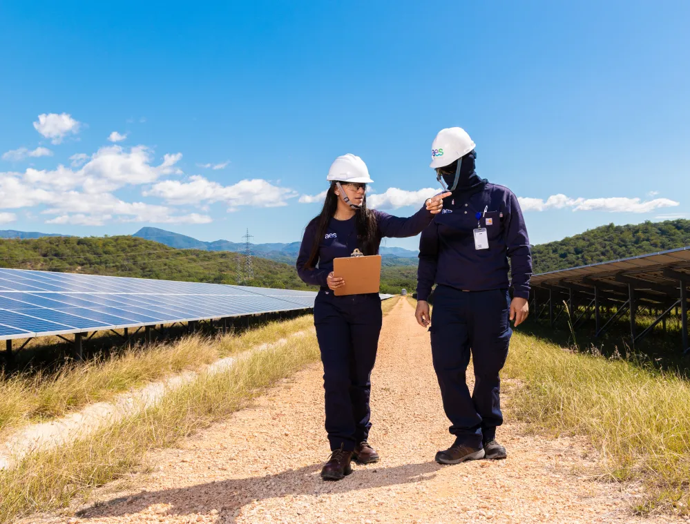 Two people wearing hard hats and protective clothing walk along a path between rows of solar panels in a sunny landscape. One person holds a clipboard and gestures while the other listens. The background features green hills and a clear blue sky.