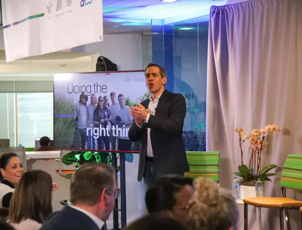 A man in a suit claps his hands while standing in front of a screen displaying the text 'Doing the right thing' with a group photo. Audience members are seated and clapping. The room is decorated with a banner, plants, and a curtain.