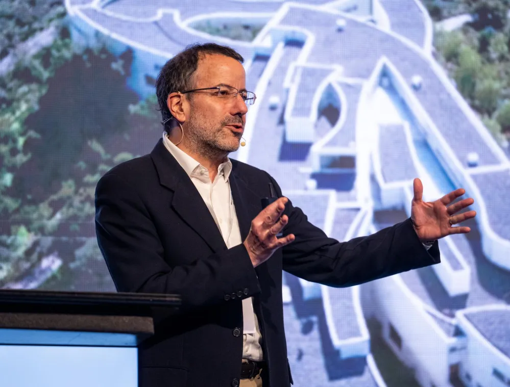 A man in a suit speaks on stage with a headset microphone, gesturing with his hands. Behind him is a large screen displaying an aerial view of a modern architectural building with curved pathways and greenery.