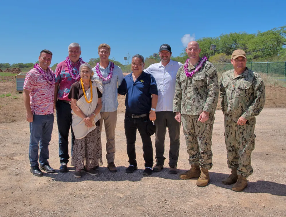 A group of eight people stands outdoors on a sunny day. Some are wearing military uniforms, while others wear casual or Hawaiian-style clothing with leis around their necks. They are standing on a dirt path with trees and a clear blue sky in the background.