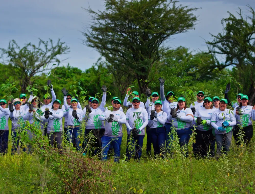 A group of people wearing matching white shirts and green caps stand in a grassy area, holding young plants. They are smiling and raising their hands, suggesting participation in a tree planting event. The background features lush green trees and a cloudy sky.