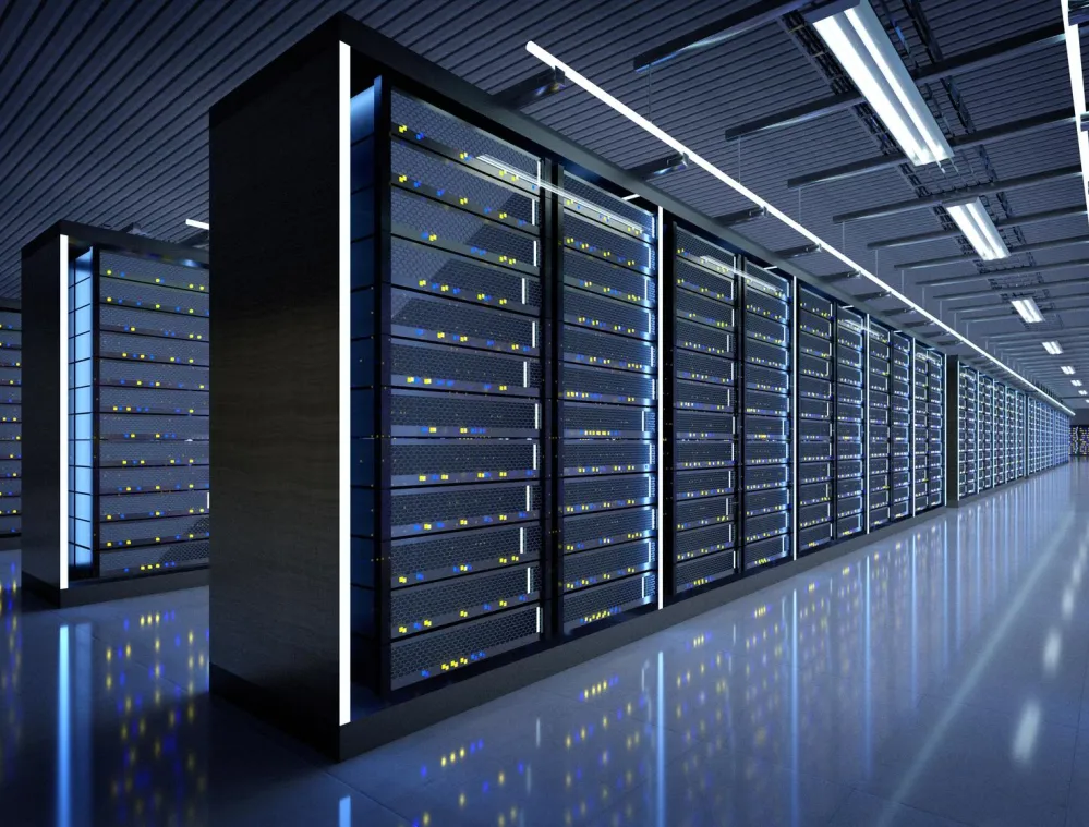 A spacious data center with rows of server racks illuminated by blue and white lights. The reflective floor enhances the lighting effect, and overhead fluorescent lights run parallel to the racks.
