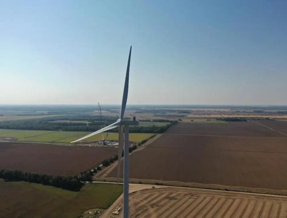 Aerial view of a wind turbine in a vast agricultural landscape with fields and distant trees under a clear blue sky.