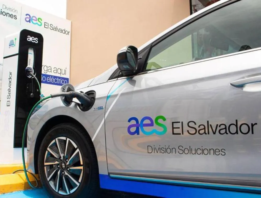 A silver electric car with the AES El Salvador logo is connected to a charging station. The car is parked on a yellow and blue painted area. A sign in the background reads 'Carga aquí tu auto eléctrico' and 'División Soluciones'.