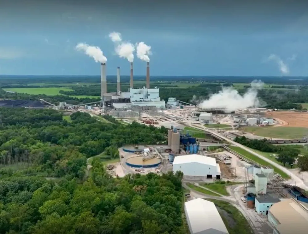 Aerial view of a power plant with four tall smokestacks emitting white smoke, surrounded by green fields and forests under a cloudy sky.
