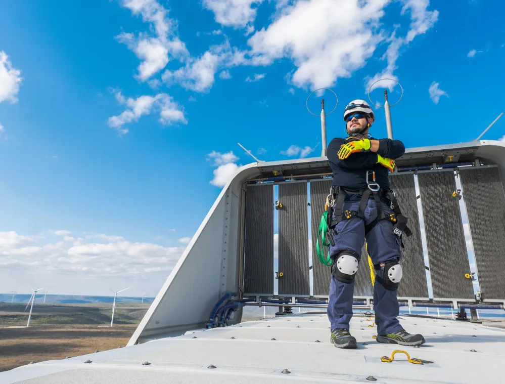 A worker wearing safety gear, including a helmet and harness, stands confidently on top of a wind turbine with arms crossed. The sky is clear with some clouds, and several wind turbines are visible in the background.