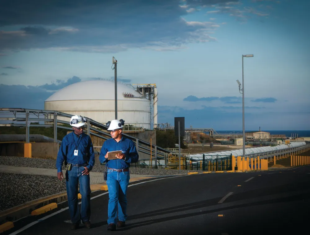Two workers wearing blue uniforms and white hard hats walk along a road in an industrial area. One holds a clipboard. Behind them is a large white storage tank and various industrial structures. The sky is partly cloudy.