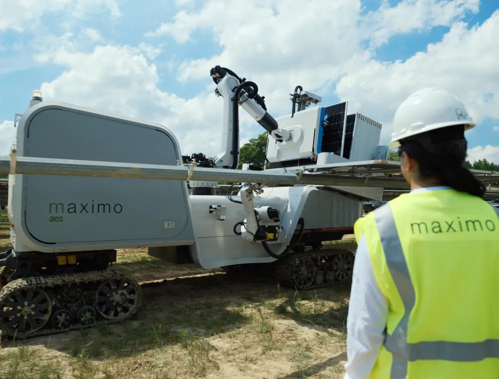 A person wearing a yellow safety vest and white hard hat stands in front of a large robotic machine labeled 'maximo aes' on a tracked base. The machine is equipped with a robotic arm and solar panels, set in an outdoor environment with a partly cloudy sky.
