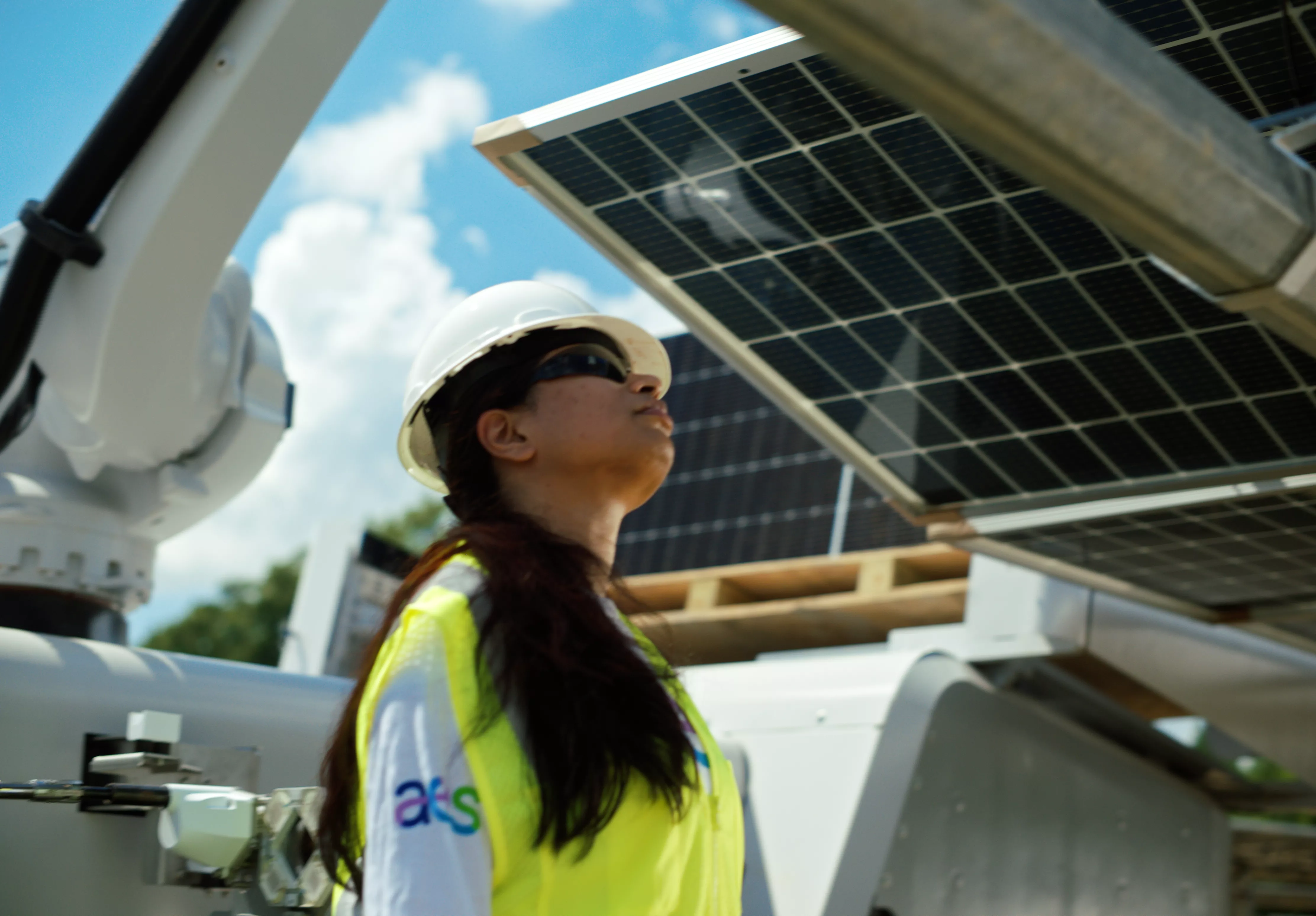 A person wearing a white hard hat, safety glasses, and a yellow safety vest is looking up at a solar panel. The person is standing outdoors with a partly cloudy sky in the background. Industrial equipment is visible around them.