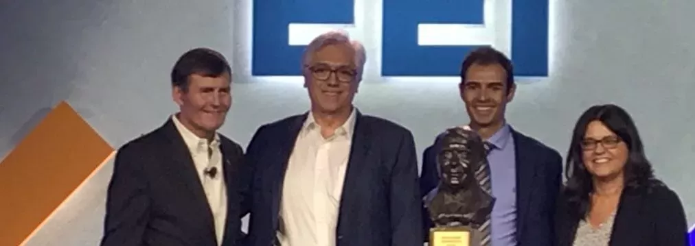 Four people stand together on a stage, smiling. One person holds a bust trophy. The background features a large logo with blue letters.
