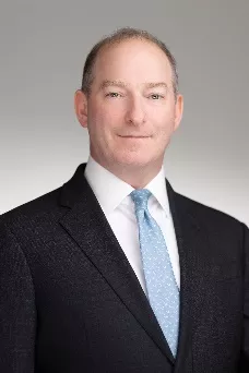 A person wearing a dark suit, white shirt, and light blue tie, standing against a plain gray background.