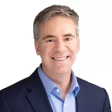 A man with short gray hair wearing a blue suit jacket and light blue shirt smiles against a white background.
