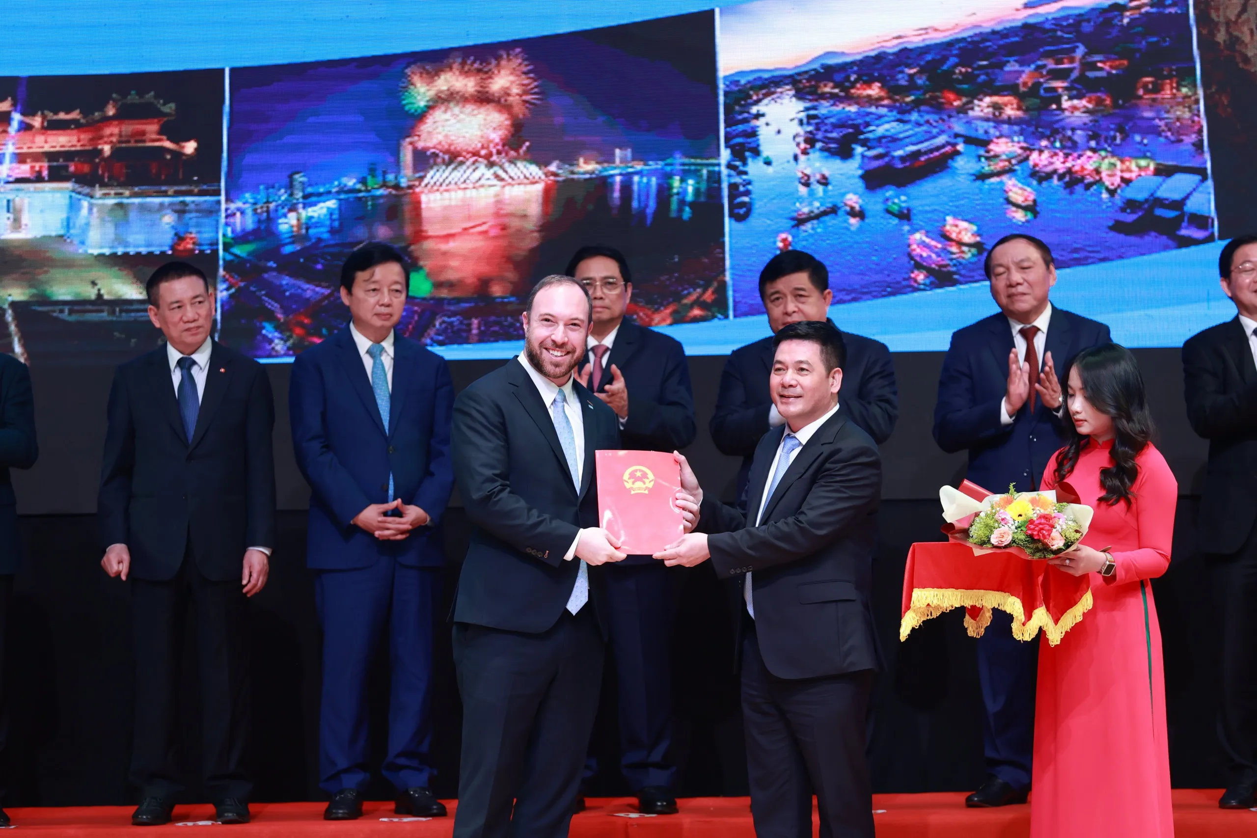 Two men in suits stand on a stage, holding a red document together. Behind them, several other people in suits are clapping. A woman in a red dress holds a floral arrangement on a tray. The background features images of colorful cityscapes and fireworks.