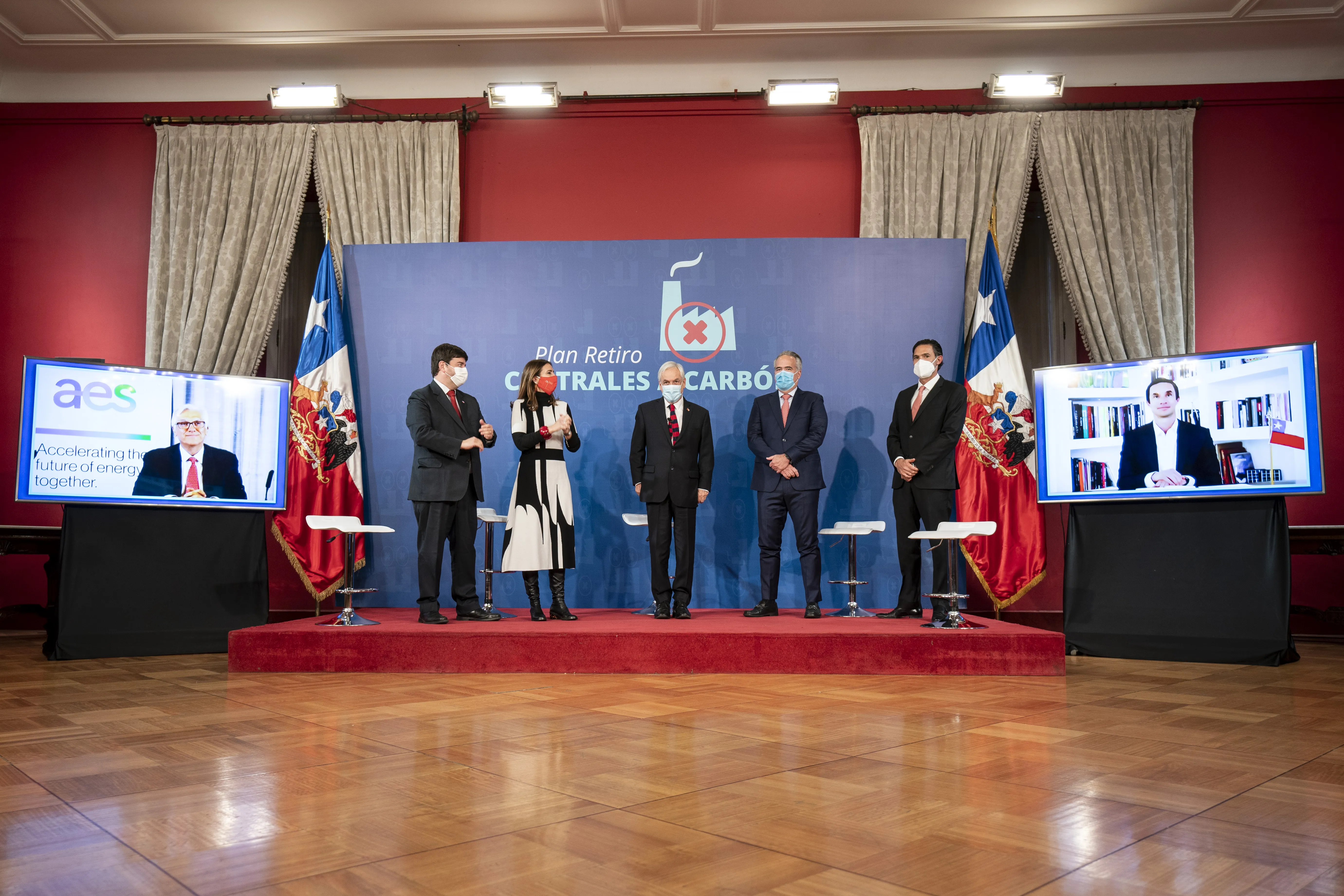 Five people wearing masks stand on a stage in front of a backdrop with the text 'Plan Retiro Centrales Carbón.' Two screens on either side display virtual participants. The setting is formal, with flags and curtains in the background.