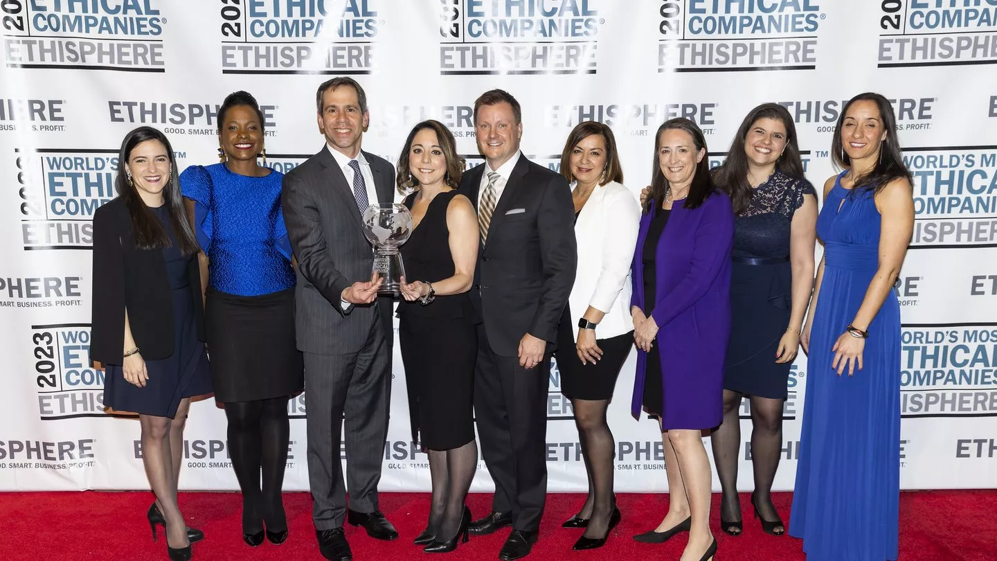 A group of nine people, dressed in formal attire, stand on a red carpet holding a trophy. They are smiling in front of a backdrop with the text 'World's Most Ethical Companies 2023' and 'Ethisphere'.