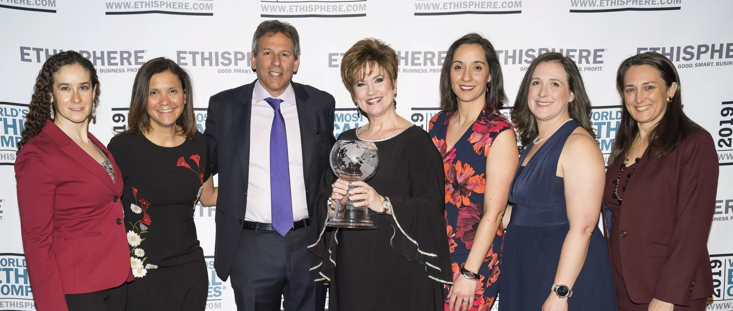 Seven people posing together at an event in front of a backdrop with 'Ethisphere' logos. One person in the center is holding a glass award shaped like a globe.