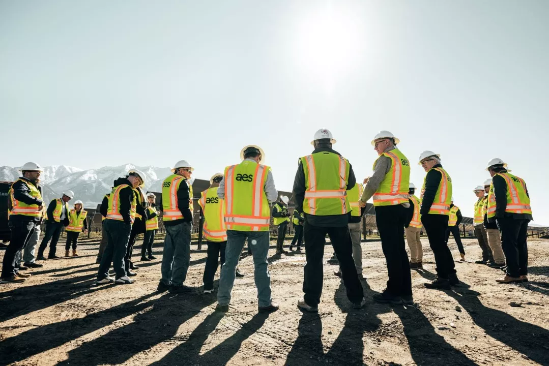 A group of people wearing hard hats and high-visibility vests stand in a circle on a construction site with mountains in the background. The sun shines brightly in the clear sky.