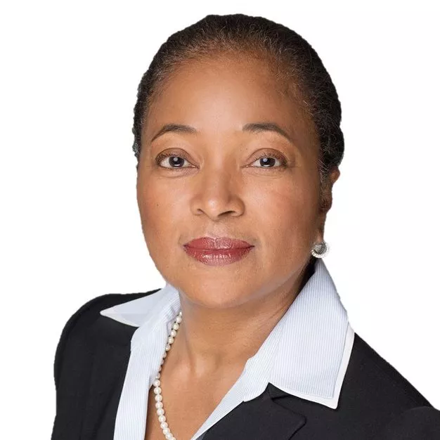 Portrait of a person with short hair, wearing a black blazer, white collared shirt, pearl necklace, and earrings, looking directly at the camera.