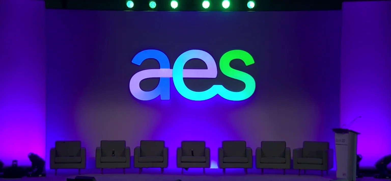 A stage setup with the large letters 'aes' illuminated in blue and green against a purple backdrop. Six empty gray chairs are lined up in front of the backdrop, and a podium with a microphone is on the right side.