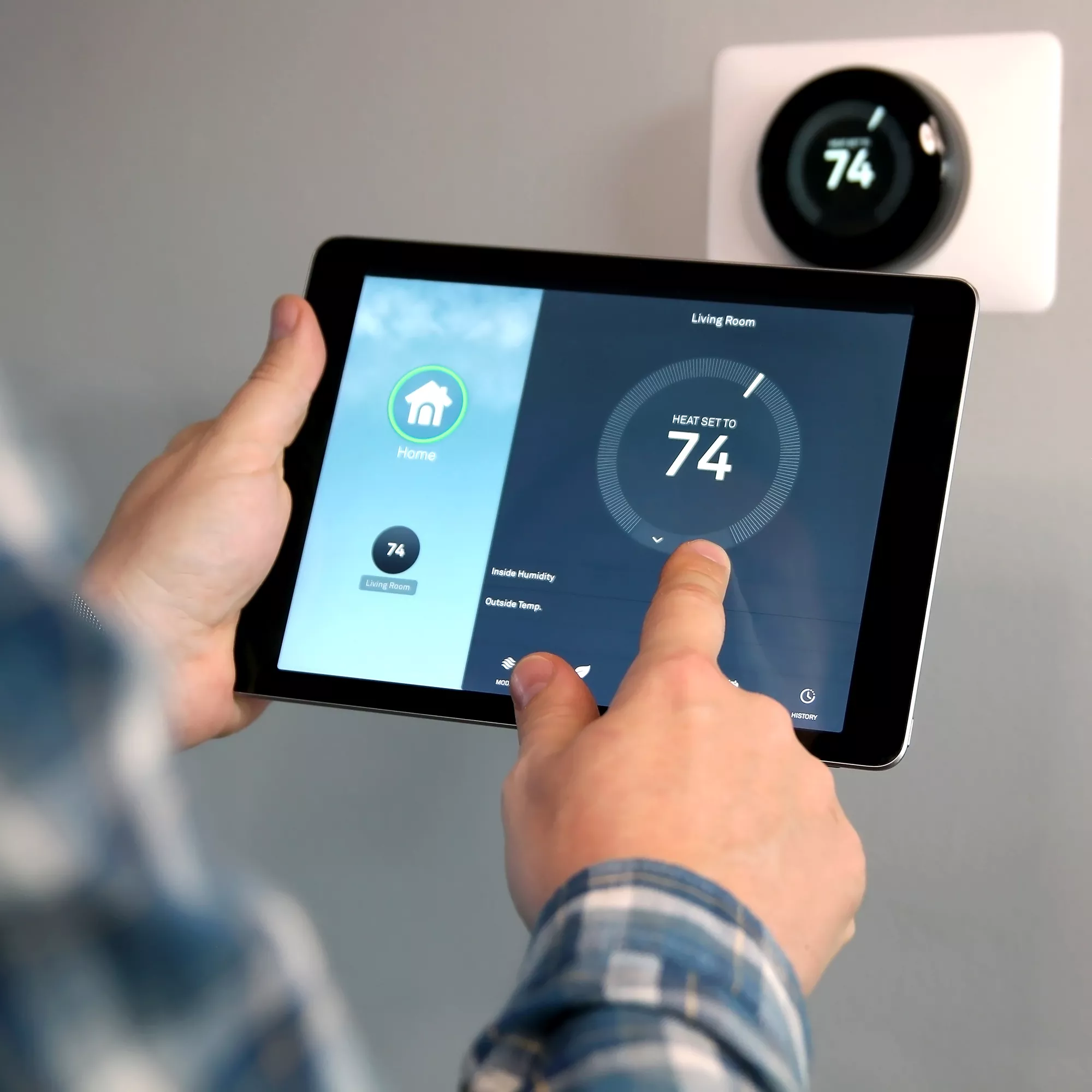 A person adjusts a smart thermostat using a tablet. The tablet screen shows a home automation app with the temperature set to 74 degrees for the living room. A wall-mounted thermostat displays the same temperature.