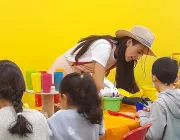 A woman wearing a hat and apron helps children with a craft activity at a table. The table is covered with an orange cloth and has colorful cups and craft supplies. The background is a bright yellow wall.