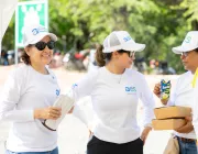 Three people wearing white shirts and caps with 'aes' logos are standing outdoors. One person is holding a pair of gloves, another is holding a juice box, and the third is holding two cardboard boxes. They appear to be engaged in conversation. Trees and a green sign are visible in the background.