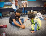 A group of children and a teenager are seated on the floor engaged in a hands-on activity with spaghetti and marshmallows. The teenager assists a child wearing a yellow vest. In the background, a woman stands nearby, and a table with various items is visible.
