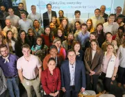 A large group of people smiling and looking up at the camera. They are gathered indoors with a banner in the background that reads, 'Safety Day, every day: Putting Gen AI to work for Safety.' The group is diverse, with a mix of men and women, and they appear to be attending a corporate event or meeting.