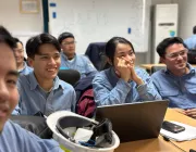 A group of young adults in a classroom setting, wearing blue shirts, attentively listening. A woman sits with hands on her chin, next to a man smiling. A hard hat and a laptop are on the desk in front of them.